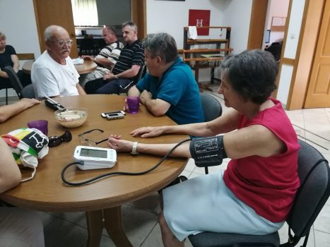 Kontrola zdravstvenog stanja, na fotografiji skupina ljudi sjedi za dva stola, u prvom planu je žena koja mjeri razinu krvnog tlaka