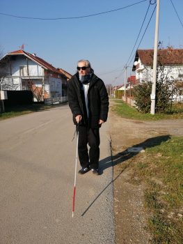 Na fotografiji se nalazi muška osoba koja se kreće ulicom uz pomoć bijelog štapa.