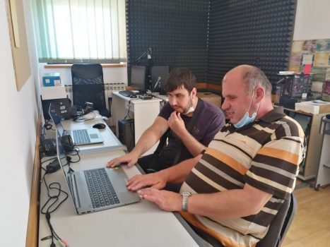 Na fotografiji se nalaze dvije muške osobe. Jedna muška osoba pokazuje nešto na lyaptopu drugoj muškoj osobi u sklopu informatičke radionice.