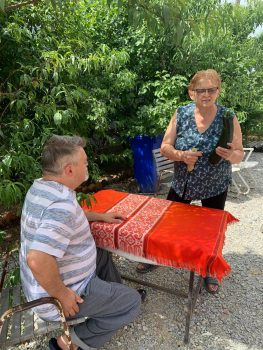 Na fotografiji se nalaze muška i ženska osoba. Muška osoba sjedi u dvorištu za stolom, a ženska osoba stoji pored stola.