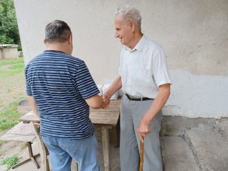 Na fotografiji se nalaze dvije muške osobe koje se rukuju i razgovaraju pored kuće.