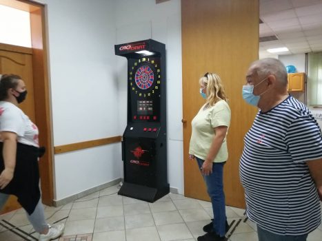 Na fotografiji se nalaze jedna mlađa ženska osoba i dvije starije muška i ženska osoba koji igraju pikado