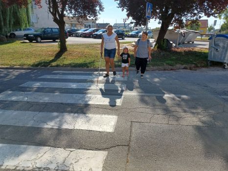 Na fotografiji se nalazi ženska osoba koja sa bijelim štapom u jednoj ruci i djetetom u drugoj prelazi kolnik na zebri uz nadzor instruktora orijentacije i kretanja.