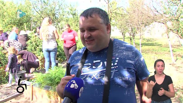 Na fotografiji se nalazi muška osoba koja daje intervju novinarima, a skupina ljudi bere rajčicu na povišenim vrtnim gredicama.