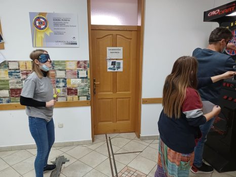 Na fotografiji se nalaze dvije mlađe ženske osobe i jedna muška osoba Muška osoba vadi strelice iz pikada dok mu ženska osoba pomaže, a druga ženska osoba sa povezom na očima čeka svoj red za bacanje strelica.