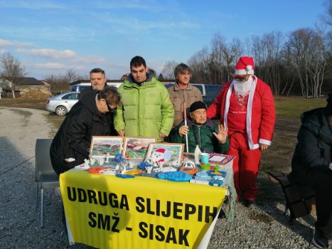 Na fotografiji se nalazi Božićni štand Udruge slijepih SMŽ - Sisak i njeni članovi pored štanda u društvu Djeda Mraza.