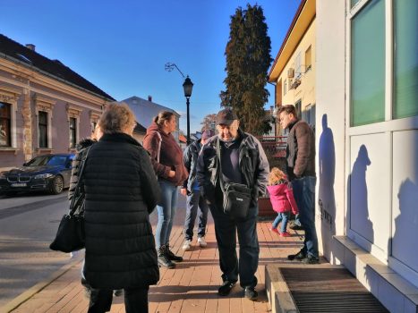 Na fotografiji se nalazi skupina ljudi, koja stoji na pločniku ispred kuće.